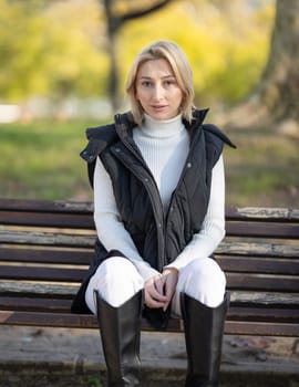 Blond fashionable woman wearing turtleneck, bodywarmer and boots enjoying sunny autumn day in park