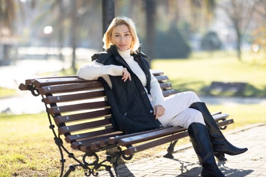 Blond woman wearing black vest and boots enjoying sunny autumn day in park