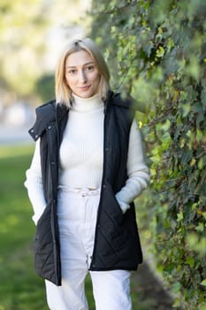 Young blonde woman wearing black vest and turtleneck white sweater posing in a park