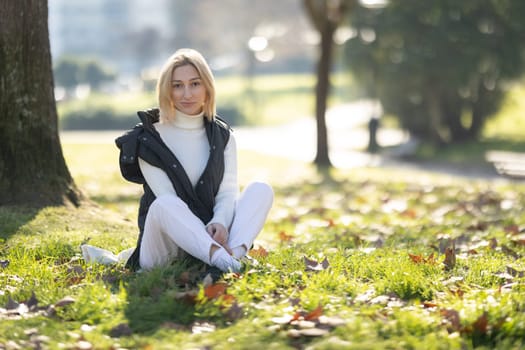 Young woman enjoying a sunny autumn day in a city park, sitting on the grass and relaxing