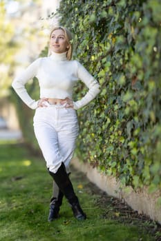 Young blond woman wearing white turtleneck sweater, white pants and black boots posing in a park with green ivy wall in the background