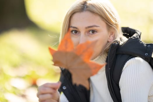 Young woman holding a large orange leaf in front of her face in a park during autumn