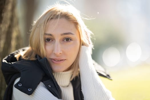 Young blonde woman wearing a white turtleneck sweater and a black vest, touching her hair while posing outdoors in a park during winter