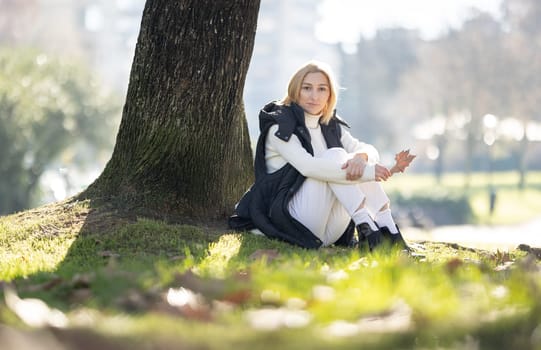 Young woman enjoying a sunny autumn day in a city park, holding a dry leaf and sitting under a tree