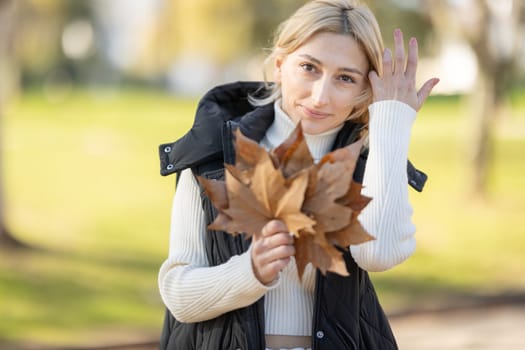 Woman wearing turtleneck and vest holding dry leaves in a park during autumn and fixing her hair