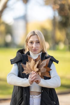 Blond woman wearing turtleneck sweater and vest holding dry leaves in a park during autumn