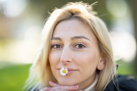 Portrait of a smiling blonde woman holding a small daisy flower in her mouth, enjoying a sunny day at the park