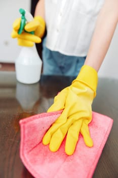 Woman cleaning table with rag and cleaning solution bottle on top, wearing yellow gloves.