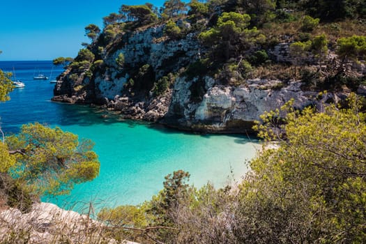 Cala Macarella in Menorca, Balearic Islands, Spain, from the mountain.Turquoise and calm waters, bright blue cloudless sky, no people, pine trees, cliffs, rocks, white sand, sailboats.