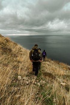 Three hikers descend a grassy slope toward the ocean, surrounded by rugged cliffs and shifting clouds. The atmosphere feels adventurous and serene as they navigate the landscape.