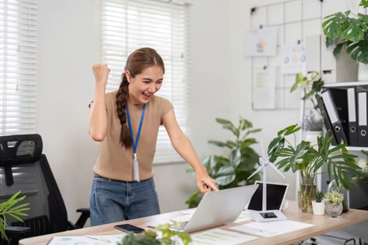 A young woman at her office desk, celebrating a recent achievement, surrounded by plants, showcasing a vibrant and sustainable work environment.