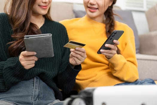Two young women enjoying a shopping moment, discussing digital payments with a credit card and a tablet in hand while sitting on a cozy couch.
