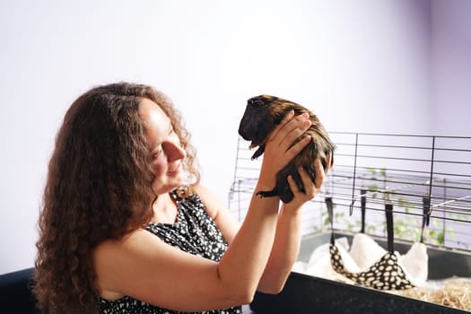 A woman holds a guinea pig close to her face, smiling with joy. The room features a guinea pig cage nearby, indicating a warm, inviting home environment.