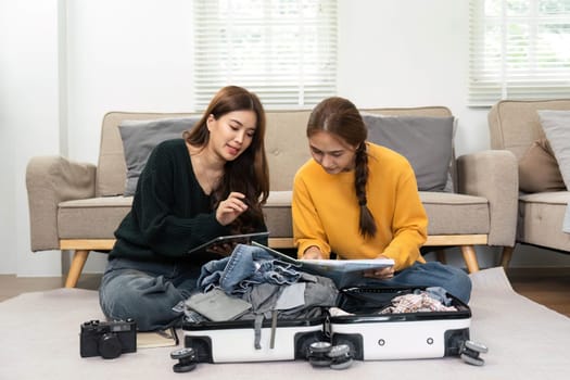 Two women getting ready for a vacation by packing clothes into a suitcase while discussing plans on a digital tablet.