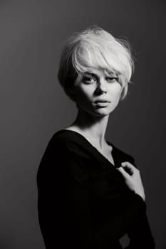 A striking black and white image featuring a young woman with short, tousled blonde hair and a black off-shoulder top. Her thoughtful expression contrasts with the soft neutral background, creating a