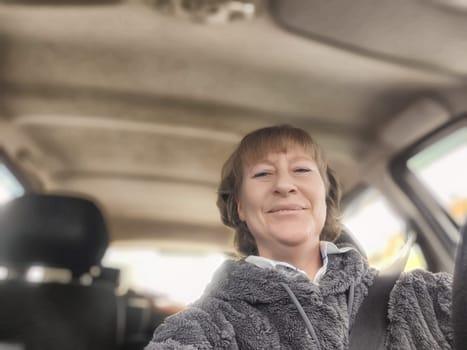 A middle-aged woman captures a cheerful moment while driving in her car, dressed comfortably in a soft hoodie.