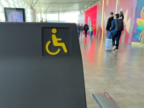 Accessible seat marked with yellow wheelchair icon in public indoor space, with people walking in background, inclusive travel and accessibility in transportation hubs