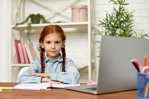 happy redhead little child girl using laptop for studying online learning system. online lesson at home. girl looking at the camera