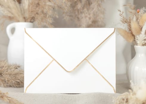 Golden edge envelope against dry beige pampas grass bouquets on linen tablecloth closeup, mockup. Boho scene with blank envelope, Bohemian or close to nature wedding stationery