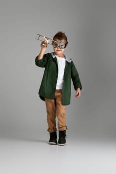 happy child boy in retro glasses playing with wooden toy airplane on studio background. dream of becoming a pilot
