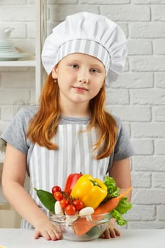 little child girl in chef hat and an apron holding bowl of fresh vegetables