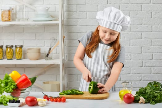 little girl in chef hat and an apron cutting green pepper in the kitchen. child cooking side dish