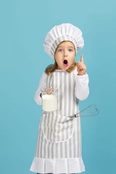 surprised little girl in chef hat and apron with flour in measuring cup and whisk pionting up on blue background. child dreams of becoming a chef. surprised expression. new idea