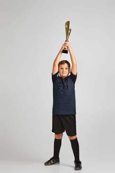 Football player holding a golden cup. little child boy playing soccer and holding trophy on gray background