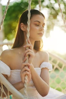 A serene young woman in a white dress, reflecting peace while sitting on a swing, illuminated by golden sunlight in a vibrant green setting, ideal for wellness themes