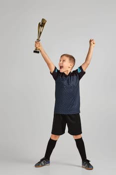 Football player holding a golden cup. little child boy playing soccer and holding trophy on gray background. kid celebrating success