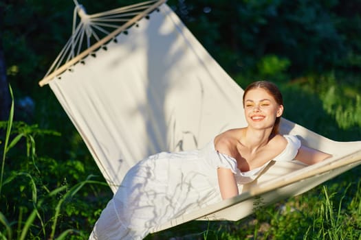 A cheerful young woman in a white dress enjoys a tranquil moment in a hammock, surrounded by lush greenery, basking in warm sunlight, conveying relaxation and happiness