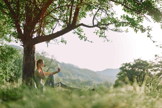 A young woman sits gracefully under a tree, sipping a refreshing drink, surrounded by lush greenery The scene reflects serenity and a healthy lifestyle in nature