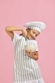 smiling little girl in chef hat and apron mixing something with a whisk in bowl over pink background. child dreams of becoming a chef