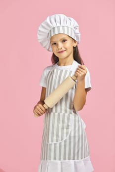 cute little girl in chef hat and apron holding rolling pin on pink background. child dreams of becoming a chef