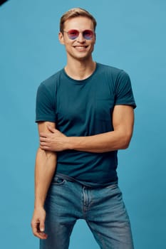 smiling young man wearing sunglasses and casual t-shirt standing relaxed with crossed arms against blue background, showing positive emotion and modern style. Concept: mental health and gadgets.