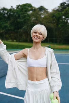Energetic young woman with short blonde hair wearing a white sports top and skirt, posing joyfully on a tennis court, representing fitness and a healthy lifestyle amid a vibrant green backdrop