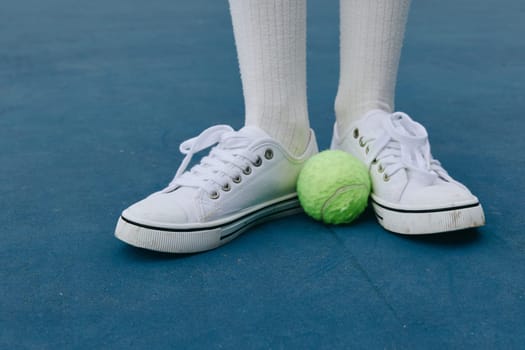 A young athlete positions themselves on a blue tennis court, standing with white sneakers next to a vibrant green tennis ball, embodying an active and healthy lifestyle in sports