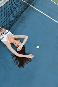 A serene young woman in a white outfit lies on a blue tennis court, symbolizing a sports lifestyle and emphasizing relaxation and wellness in sports activities