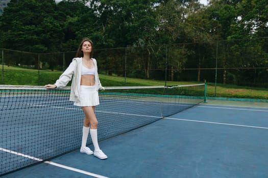Confident young woman in white sports attire poses by the tennis net, showcasing a healthy lifestyle and sports enthusiasm, ideal for fitness and sporting themes