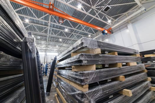 Stacks of metal profiles wrapped in plastic film are stored on wooden pallets inside a spacious warehouse, ready for industrial use