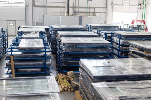 Stacks of pvc windows wrapped in stretch film, stored on metal racks and wooden pallets in a factory warehouse, ready for shipping