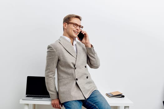 Confident smiling man dressed in smart casual blazer appears engaged in phone conversation, standing in a minimalistic office setting with a laptop and notepads on the desk, bright and professional atmosphere. Concept: business communication, workplace.