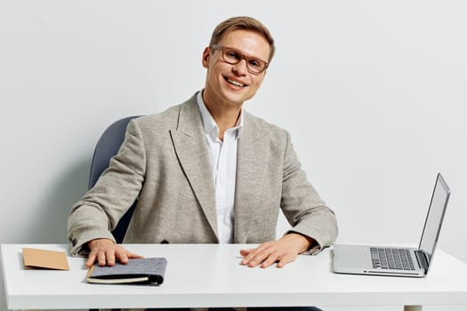 Friendly man in formal attire working at desk in bright office environment with laptop, notebook, and stationery, smiling warmly at camera. Positive attitude, professional vibe, workspace scene. Concept: modern work