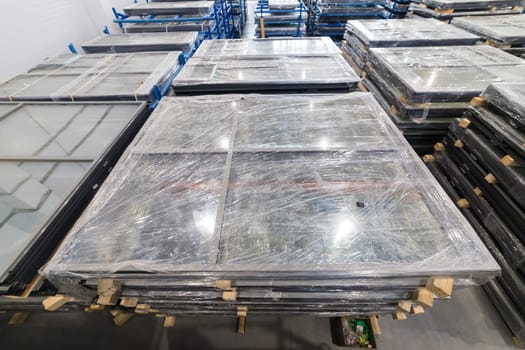 Large window frames wrapped in plastic film, stored in a warehouse, awaiting transportation to construction site or customer