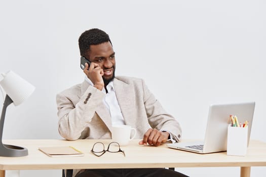 Business professional man with joyful expression speaking on cell phone while seated at desk in contemporary office space, surrounded by laptop, glasses, and paperwork, conveying communication, workplace, success, and technology concepts.