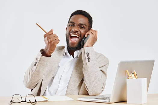 Happy man engaging in a lively conversation on mobile phone, dressed in smart casual blazer, sitting at a desk with laptop, glasses, and stationery, on plain white background. Concept: business communication and success.