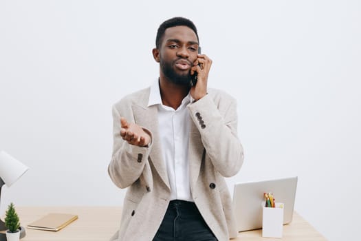 Professional man communicating via mobile phone in a contemporary office environment, dressed in a beige blazer and white shirt, gesturing with hand, seated at desk with laptop and supplies, bright clean background, concept of business conversation, modern work lifestyle.