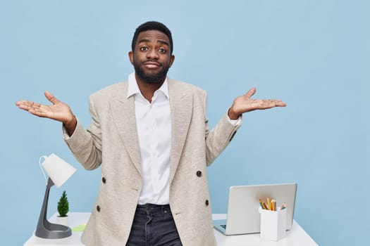 Young man with confused or unsure expression, standing at a contemporary office desk with laptop, stationery, and small plant, on solid light blue background. Casual business look, workspace setting, indoor, modern office style, professional background, everyday office scene, workplace mood.