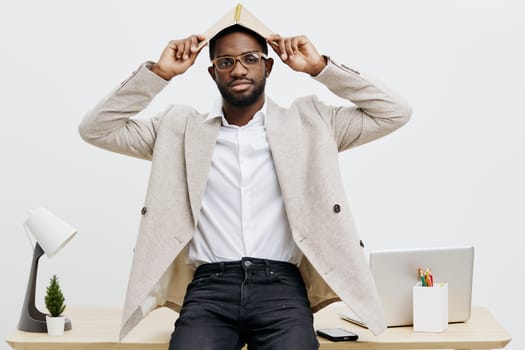 Confident young man with fashionable glasses and trendy beige blazer, sitting at a clean, modern office desk, holding a hat, expressing style and professionalism. neutral background, studio shot, contemporary fashion concept