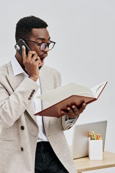 Confident young man of African ethnicity with glasses, dressed in a beige blazer and white shirt, engaged in phone conversation while holding an open notebook, standing in a clean, minimalistic office with a desk and stationery. Office worker lifestyle